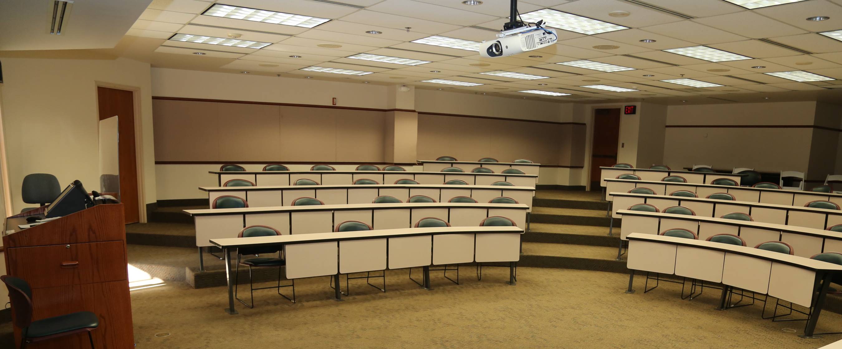 Room with tiered seating and chairs, a teachers desk with a computer, and a ceiling-mountedprojector