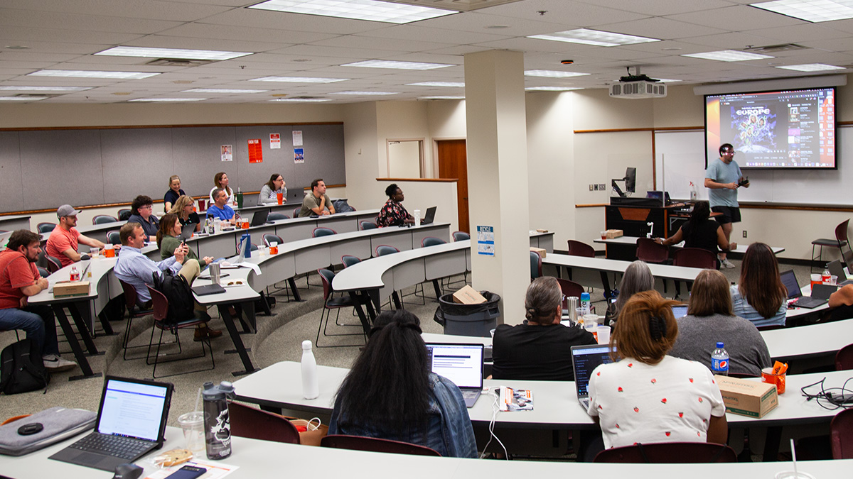 A group of people in a tiered lecture room listen to someone giving a presentation in front of a projector screen.