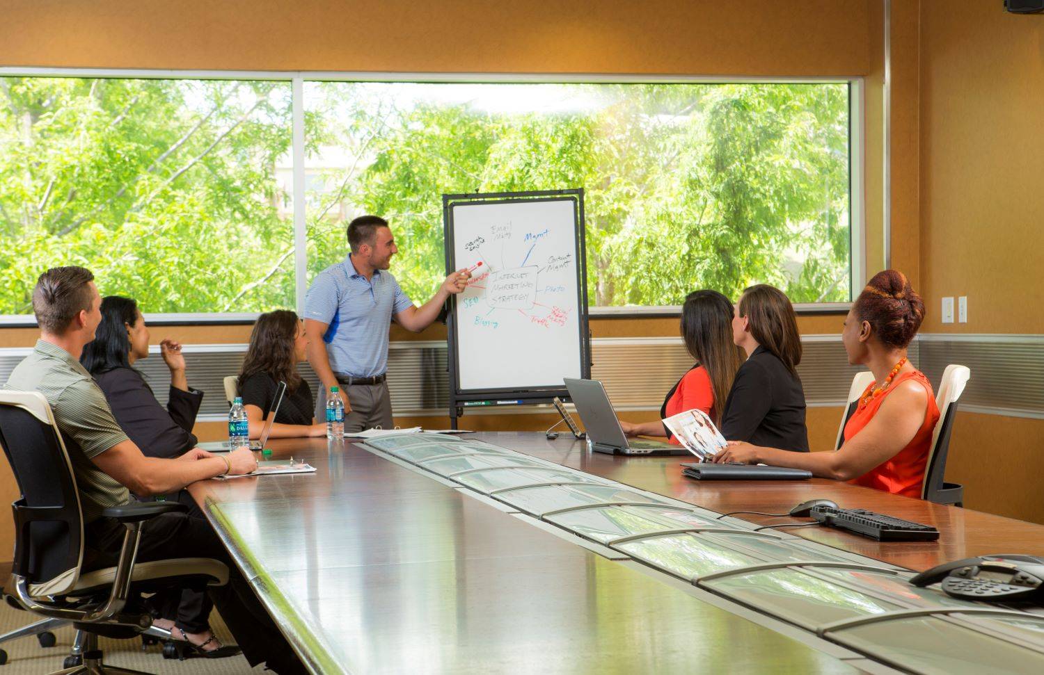 Six professionals sit around a conference table while a man points to a whiteboard with a marketing diagram.